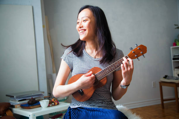 Millennial young Asian woman having fun playing Ukulele in her studio apartment