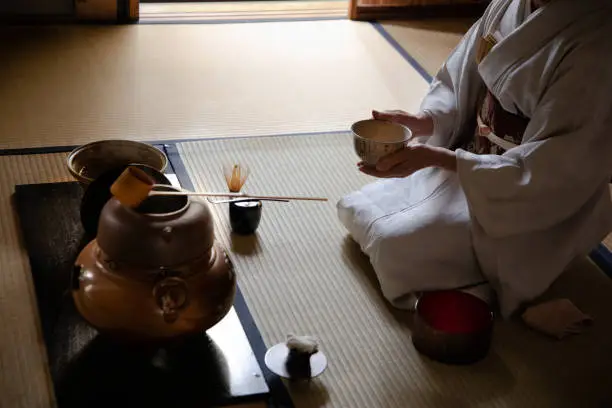A Japanese woman wearing a kimono is kneeling and preparing items for a tea ceremony. She is kneeling on a tatami mat, next to a window, holding a bowl.