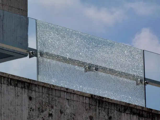 shattered glass railing panel above stone panel under blue sky and white clouds. broken laminated tempered safety glass. construction and building industry concept. 
stainless steel glass brackets.