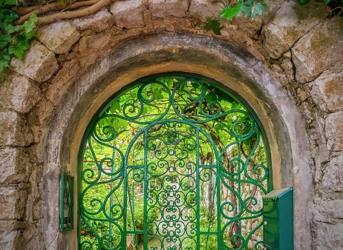 The old stone entryway is rounded on top, and covered with climbing vines. The arched, wrought iron gate is painted green and has an ornate, decorative, ornamental design. Inside the gate are a vine-covered wall, a path, and a secret garden of green plants lit by beautiful light.