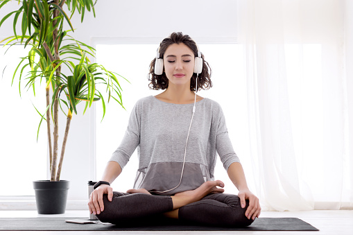 Slender female folded crossed legs in lotus position. Young athletic woman in fitness tracker and headphones is practicing yoga, meditating, relaxing while sitting in padma asana at home.