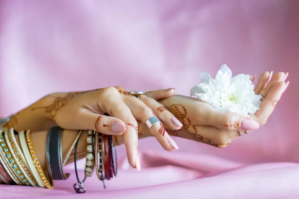 Slender elegant female wrists painted with traditional Indian oriental mehndi ornaments by henna. Hands dressed in bracelets and rings hold white flower. Light pink fabric with folds on background.