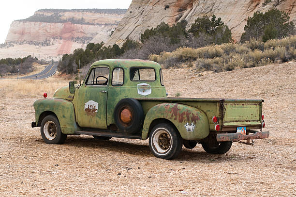The left-side rear view of an old rusty Chevrolet 3600 Pick-up covered with chipped green paint standing at the foot of a mountain. 