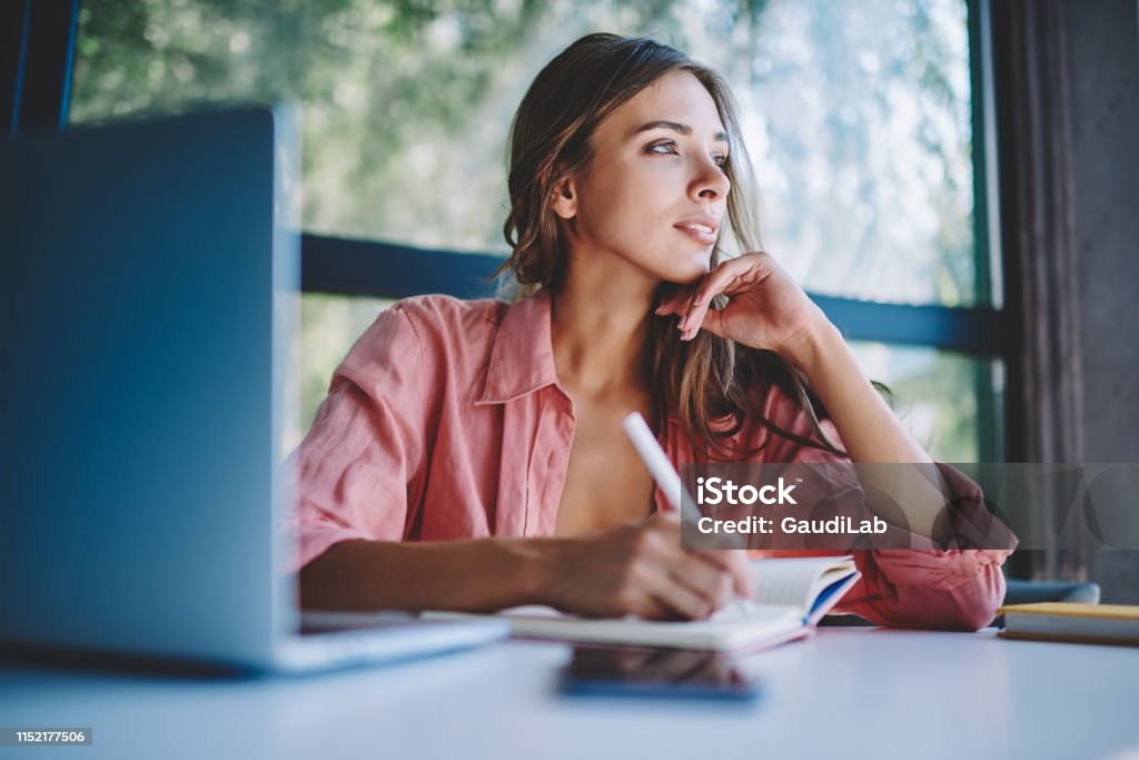 Pondering young woman looking away while making notes and thinking on creative ideas for developing own internet website.Thoughtful hipster student writing down information in notepad sitting at table One Woman Only Stock Photo