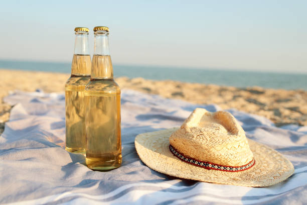 Closeup of two beer glass bottles on sandy tropical beach towel near straw hat. Blue ocean lagoon on background. Refreshing beverage on hot summer day. Two drinks near the sea shore stock photo