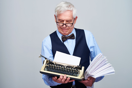 Senior elegant man holding retro typewriter and papers while contemplating on gray background