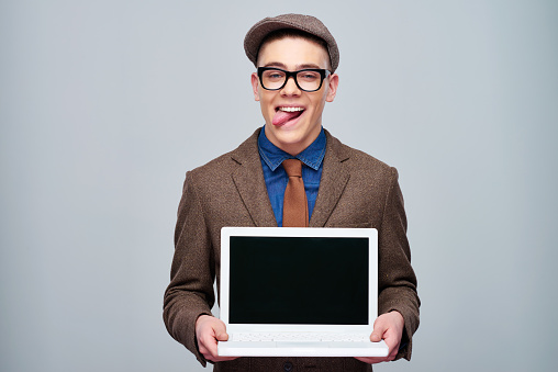 Young classy man in old-fashioned cap showing tongue while demonstrating new laptop on gray backdrop
