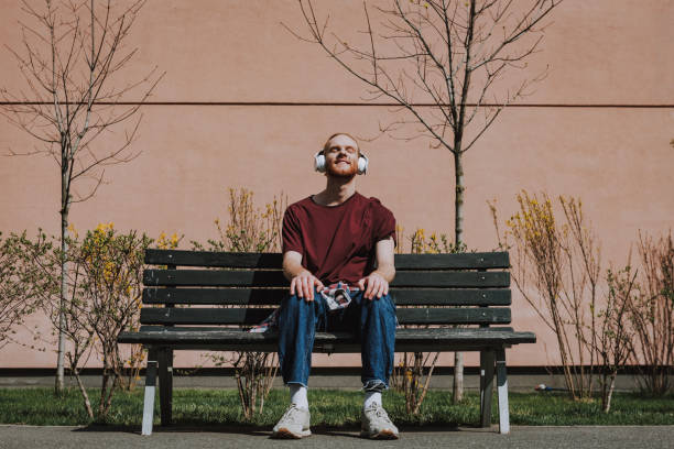Urban lifestyle concept. Full length portrait of young happy hipster guy with headphones on head closing eyes while enjoying music on bench