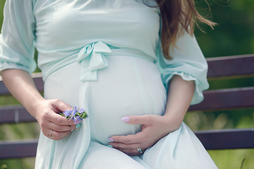 Close up of beautiful young Caucasian woman in her last trimester of pregnancy wearing a mint color vaporous summer dress and holding flowers in her hands in a caressing gesture around her baby bump.