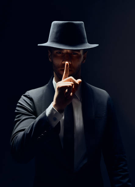 istock Serious man in black suit and hat keeps finger on lips, making hush gesture and keep conspiracy isolated on dark background 1152186190