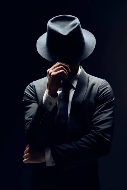 Man in suit hiding face behind his hat isolated on dark background Man in suit hiding face behind his hat isolated on dark background. secret and incognito concept mob stock pictures, royalty-free photos & images
