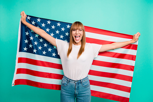 Close up photo beautiful she her lady arms hands hold spread big american flag yelling festive mood crazy carefree weekend vacation wear specs casual white t-shirt isolated teal green background.
