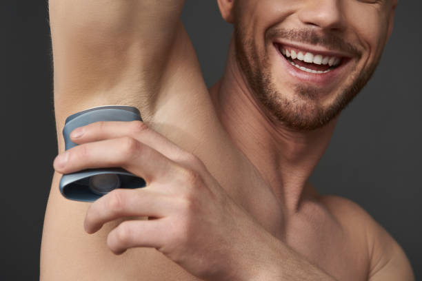 Guy takes care of his hygiene by applying deodorant to his armpit Cropped photo of young Caucasian man applying antiperspirant to his underarm. He is smiling while situating against gray background in studio. Body care and male hygiene concept deodorant stock pictures, royalty-free photos & images