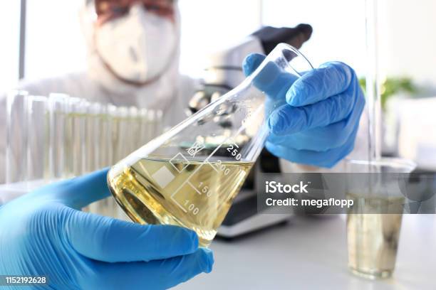 Scientist Arm In Protective Gloves Hold Yellow Liquid In Bottle Stock Photo - Download Image Now