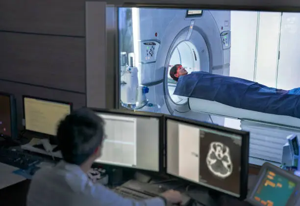 Doctor examining scan on computer while patient entering in MRI scanner. Medical expert and woman are in medical examination room. They are in hospital.