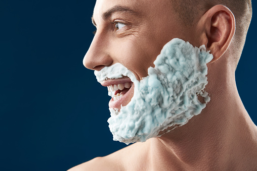 Side view of Caucasian man with foam for shaving on his face situating against blue background. Male model is smiling while looking away. Body care and male hygiene concept