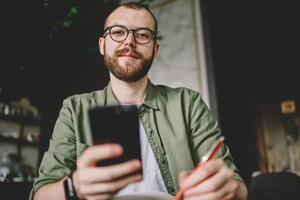 Photo of Below portrait of bearded hipster student in stylish eyeglasses for vision correction holding modern smartphone in hands and rewriting information from internet website while looking at camera