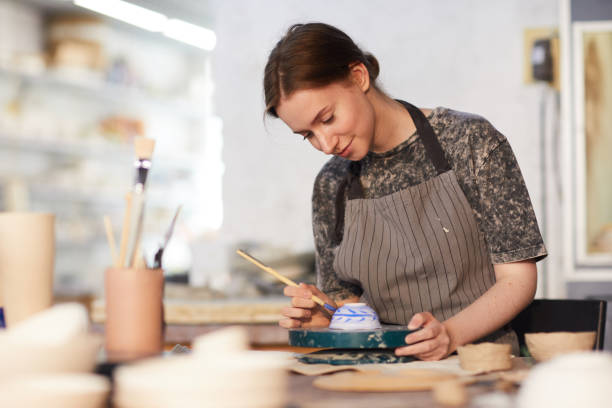 Skilled lady drawing on ceramic bowl Content attractive skilled young lady in apron standing at table and drawing blue leaves on ceramic bowl in pottery workshop pottery photos stock pictures, royalty-free photos & images