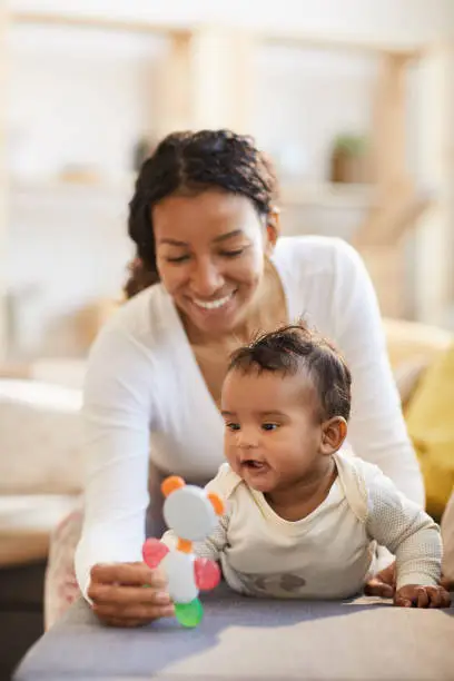 Happy young black mother showing cute toy for teething to excited baby boy on sofa, infant exploring world concept