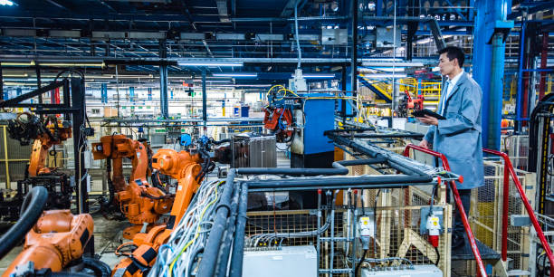 Male engineer of Asian ethnicity standing on a platform and looking at an automated production process done by robotic arms while holding a digital tablet.