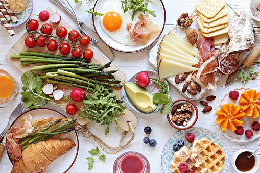 Brunch or breakfast table, meal variety with fried egg, asparagus, avocado, sausage and cheese variety, croissants, smoothie, fresh waffles and fruits. Overhead view