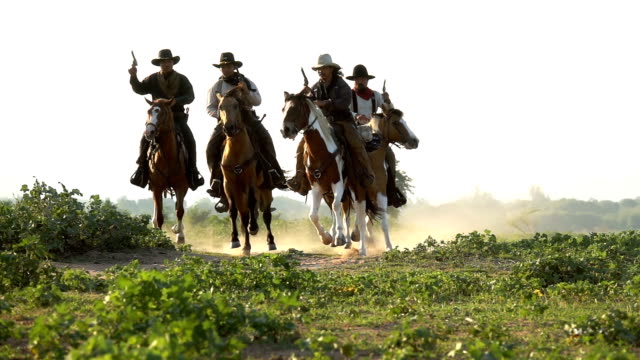 Group Of Young Cowboys Holding Gun And Riding Horse Running In The Meadow Field