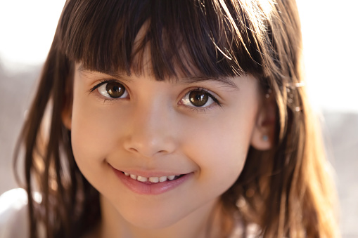 Head shot portrait of cute smiling little girl looking at camera, adorable positive kid posing at home for photo, feeling happy, cheerful funny child, model with beautiful eyes, close up
