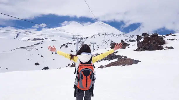 The brunette girl stands with her back with a backpack on her shoulders with her hands raised and looks towards mount Elbrus. Travelling to Russia. Freedom and joy.