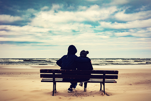 Father and daughter sitting together on a lonely bench on the beach and looking at the sea. Conceptual