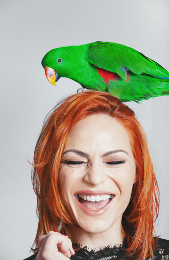 Young woman laughing with her eyes closed while green parrot is standing on her head.