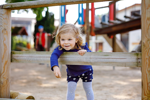 Cute toddler girl having fun on playground. Happy healthy little child climbing, swinging and sliding on different equipment. On sunny day in colorful clothes. Active outdoors game for children.
