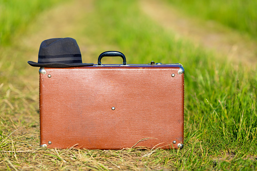 Travel and adventure concept. Vintage brown suitcase and hat on the road in the green field.