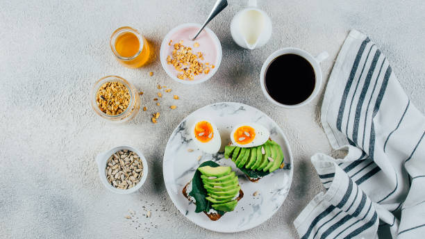 istock Healthy Breakfast concept. Sandwich with avocado, spinach, and boiled eggs, yogurt with granola, cup of coffee over concrete background, top view, banner image 1152349814
