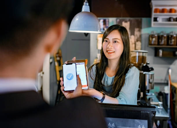 Customer paying through digital wallet at cafe Smiling female cashier holding digital tablet at checkout. Male customer is paying through smart phone. They are at cafe. asian cashier stock pictures, royalty-free photos & images
