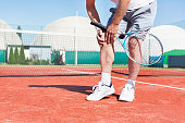 Low section of mature man holding tennis racket while suffering from knee pain on red tennis court during summer