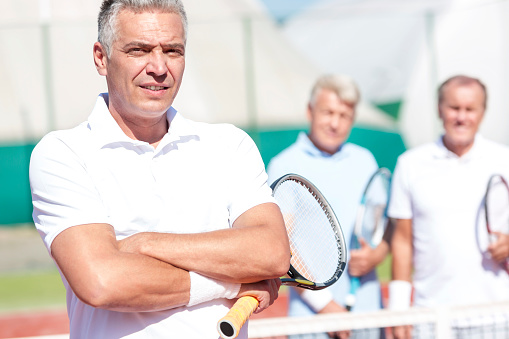 Portrait of confident mature man holding tennis racket while standing with arms crossed against friends on court during sunny day