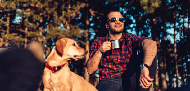 Hiker wearing plaid shirt and sunglasses resting on grass with small brown dog by the pine forest and drinking coffee during sunset