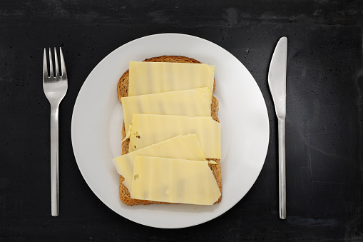 Slice of brown bread with cheese on a white plate, stainless steel silverware and black stone background.