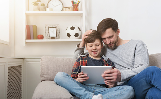 Father and son spending time together and playing on digital tablet on sofa at home