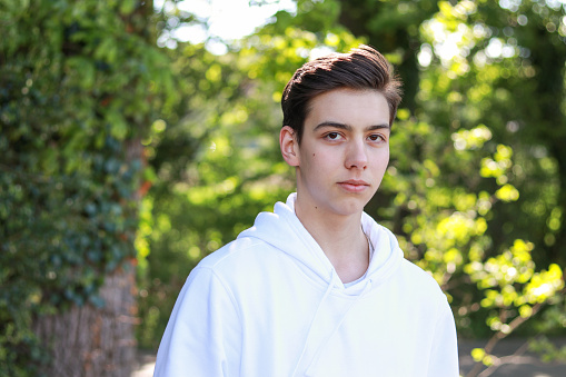 Close-up portrait of handsome serious teenager boy in white hoody in park outdoors at bright sunny day.