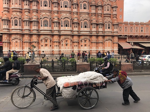 Jaipur, Rajasthan, North India - March 2, 2019:  Stock photo of loaded, bicycle trolley being pushed and pulled by local Indian men, past the Hawa Mahal built in 1799 from red and pink sandstone. This building is also known as the Palace of Winds or Palace of the Breeze.