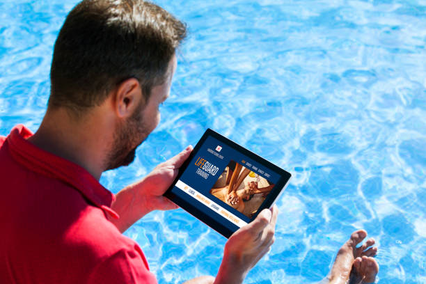 istock Man holding a tablet with emergency services training website in the screen, while sitting at the swimming pool shore. 1152396218