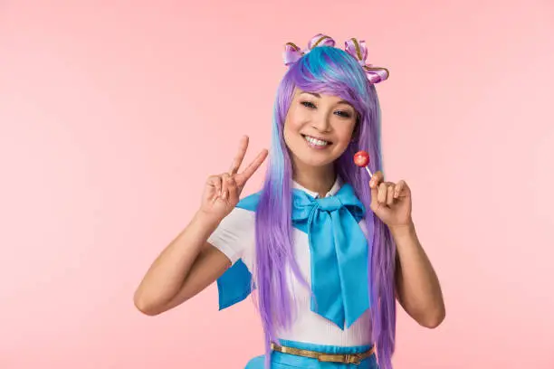 Happy asian anime girl holding lollipop and showing peace sign isolated on pink
