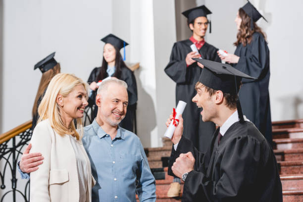 istock selective focus of cheerful parents looking at happy son in graduation cap gesturing while holding diploma near students 1152405406