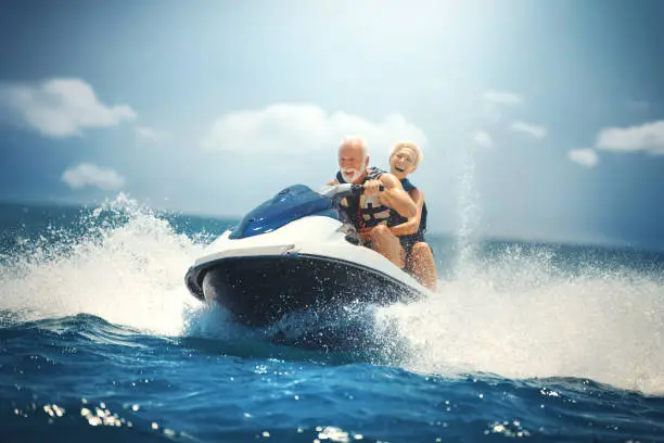 Closeup side view of a senior couple riding a jet ski on a sunny summer day at open sea. The man is driving quickly through the waves, and the lady is hardly holding on. Caught in the moment of max speed.