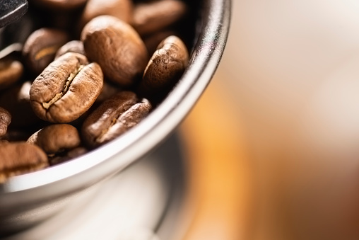 Macro image of lots of smooth brown coffee beans sitting in the top of a small manual grinding coffee machine. The focus is selective and is on the coffee bean in the lower left of the image. There is copy space to the right of the image.
