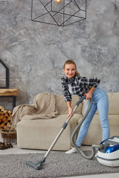 Young smiling woman vacuum cleaning the carpet in the living room, modern scandinavian interior. Busy, cleaning day. Home, housekeeping concept