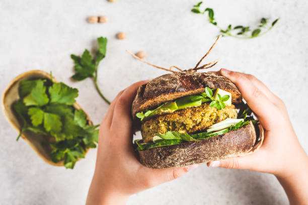 Vegan sandwich with chickpea patty, avocado, cucumber and greens in rye bread in children's hands. Vegan sandwich with chickpea patty, avocado, cucumber and greens in rye bread in children's hands, top view. veggie burger stock pictures, royalty-free photos & images