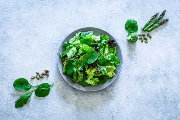 Vegan food: top view of a fresh organic green salad shot on gray abstract background. The salad is in a gray plate and fresh ingredients like broccoli, pumpkin seeds, spinach, arugula, pistachios and asparagus are out of the plate placed directly on the background. Useful copy space available for text and/or logo. Predominant colors are green and gray. Low key DSRL studio photo taken with Canon EOS 5D Mk II and Canon EF 100mm f/2.8L Macro IS USM.