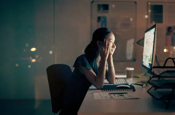 Shot of a young businesswoman looking tired while using a computer at night in a modern office